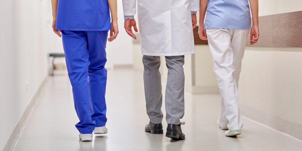 Kolme terveydenhuollon ammattilaista kävelee sairaalan käytävällä poispäin kuvaajasta.