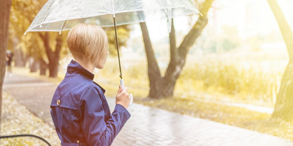 Nainen sateessa katselee puistossa kaukaisuuteen sateenvarjon alla.