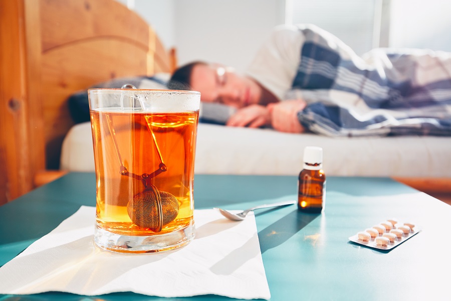 Taustalla sairaana oleva mies peiton alla sängyssä. Etualalla pöydällä kupillinen teetä ja lääkkeitä.