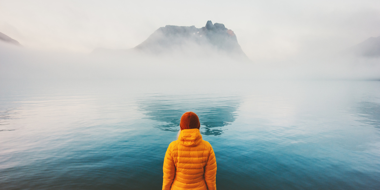 Nainen keltaisessa talvitakissa ja punainen pipo päässä seisoo ja katselee veden yli sumusta erottuvaan saareen.