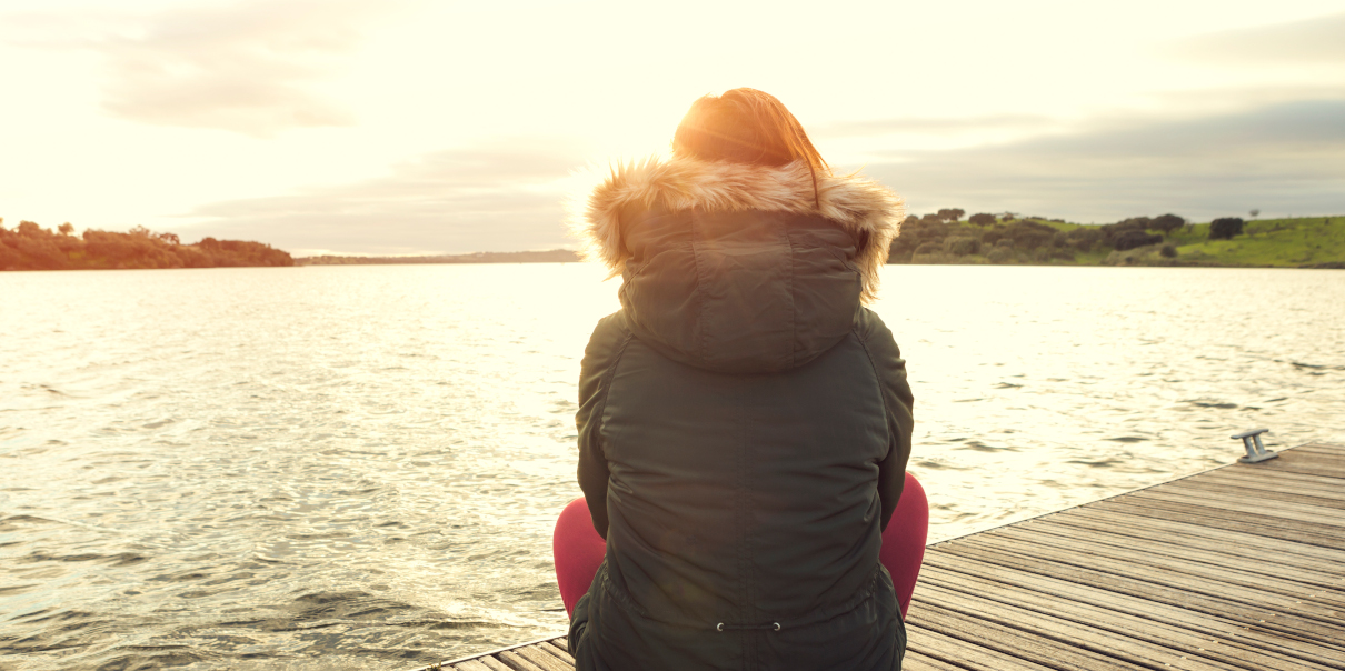 Nainen talvitakissa istuu laiturilla ja katsoo poispäin kohti aurinkoa