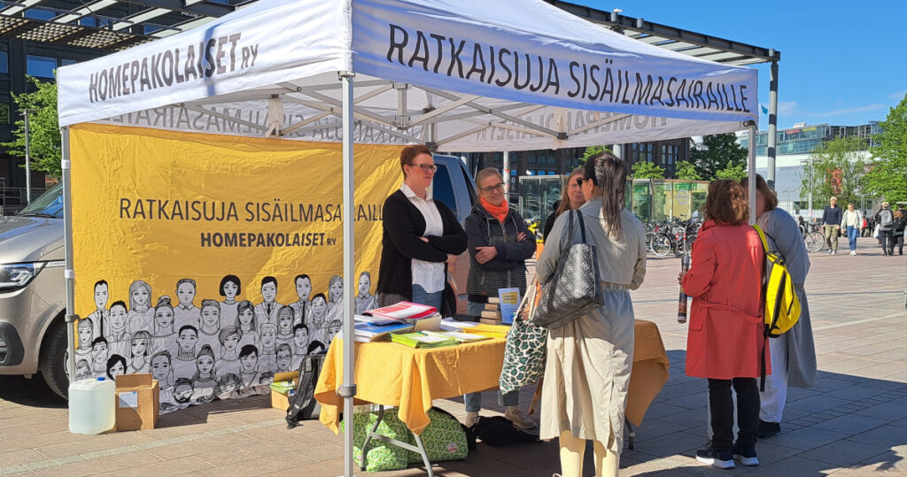 Homepakolaiset-yhdistyksen toriteltta Narinkkatorilla, teltalla esitteitä ja useita ihmisiä. Aurinko paistaa.