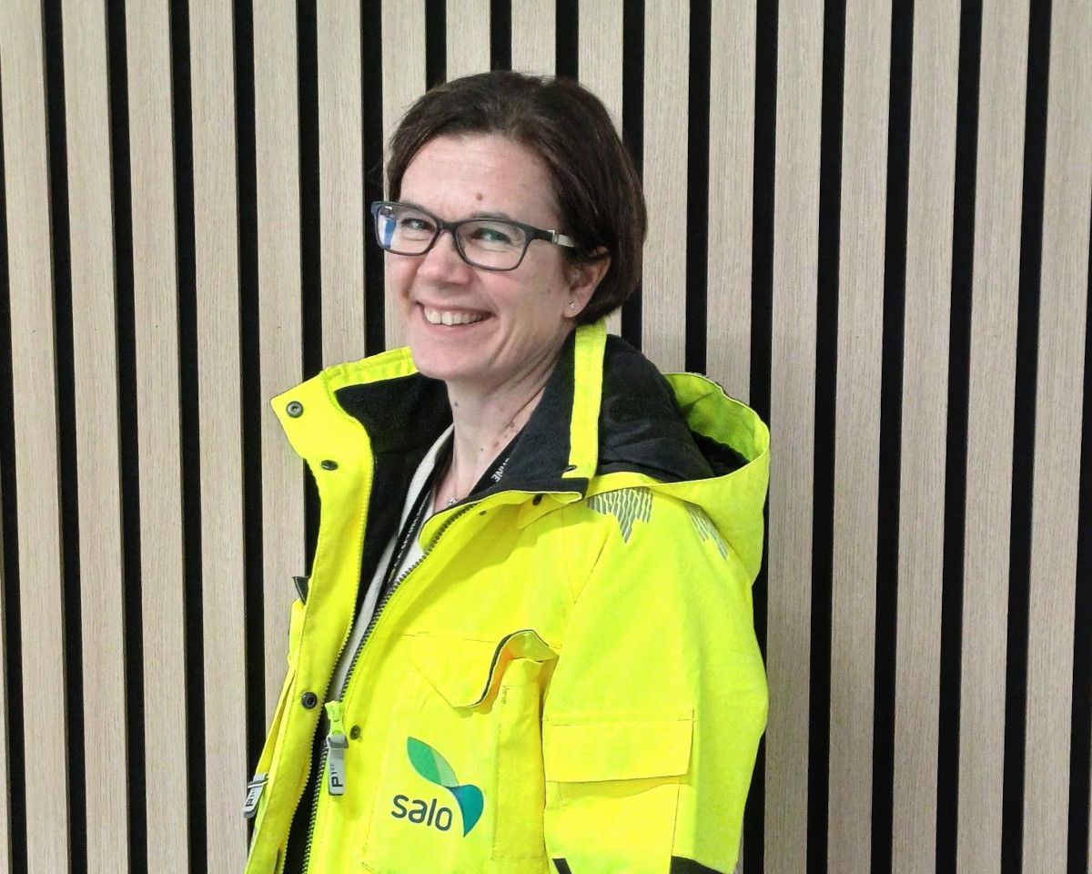 Nainen neonkeltaisessa työtakissa hymyilee ja katsoo kameraan.