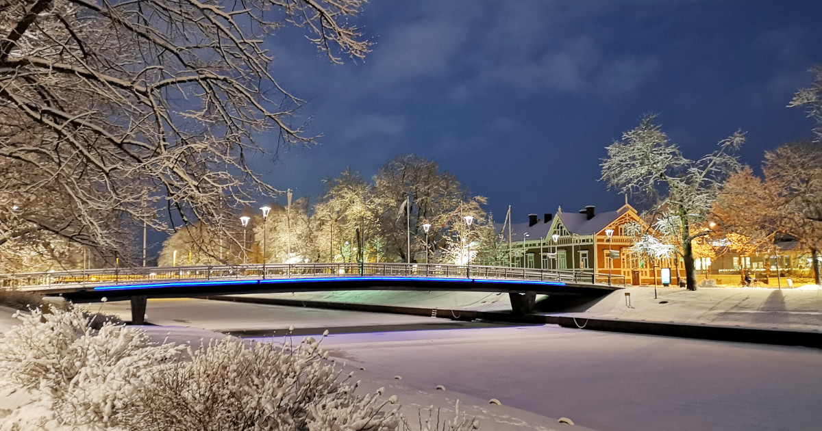 Talvimaisemassa silta kulkee joen yli, rannalla perinteinen puutalo.